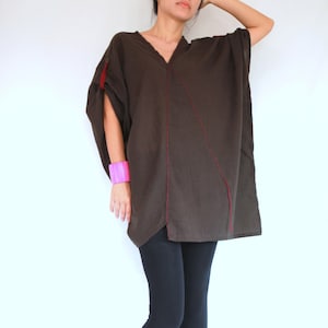 May include: A dark brown tunic with red stitching details and a V-neck. The tunic is paired with black leggings and brown wedge sandals. A bright pink bracelet is worn on the left wrist.
