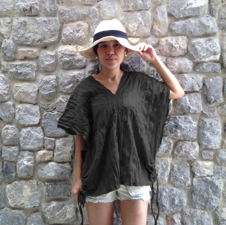 May include: A woman wearing a dark green, loose-fitting top with a v-neck and a white hat. The top has a drawstring at the waist and the woman is wearing denim shorts.