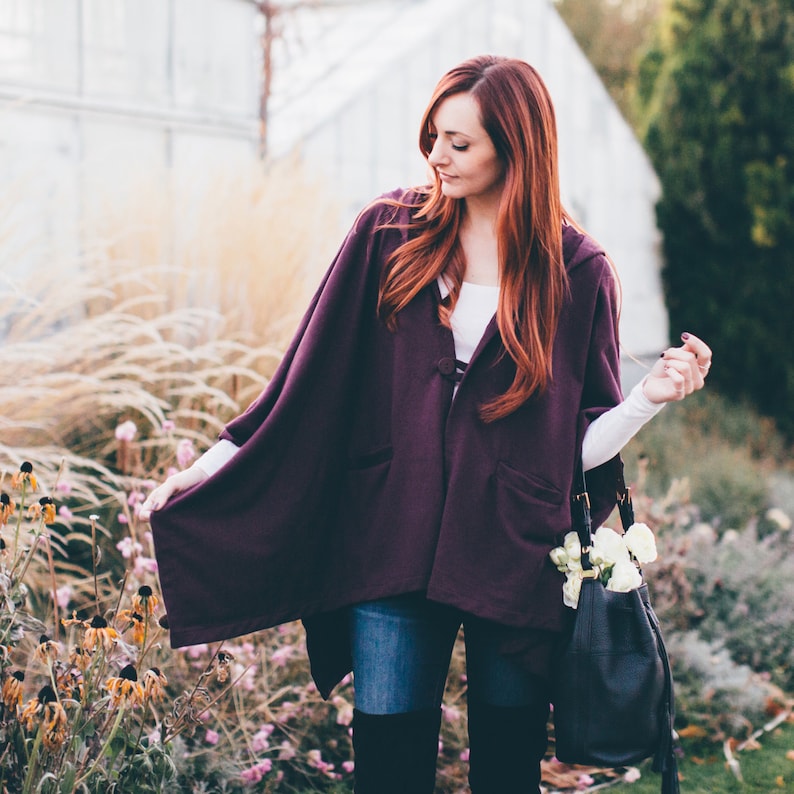 May include: A woman wearing a purple hooded cape with a white shirt underneath. She is wearing blue jeans and black over-the-knee boots. She is holding a black handbag with white flowers.