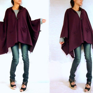 May include: A purple hooded cape with a button closure and a long, asymmetrical hemline. The cape is worn over a pair of dark blue jeans.