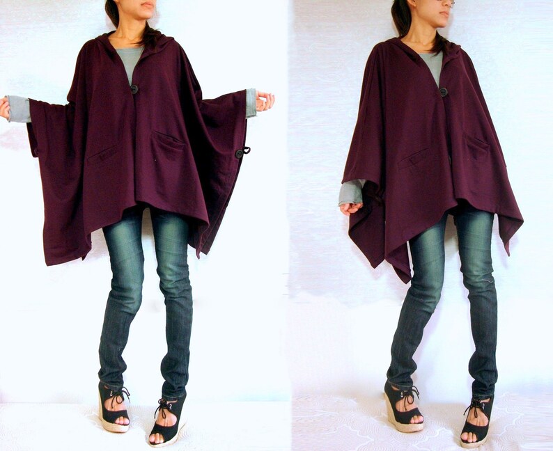 May include: A purple hooded cape with a button closure and a long, asymmetrical hemline. The cape is worn over a pair of dark blue jeans.