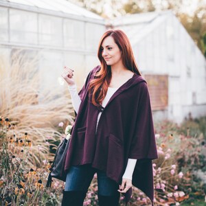 May include: A woman wearing a purple hooded cape, blue jeans, and black over-the-knee boots. She is standing in a garden with flowers and greenery.