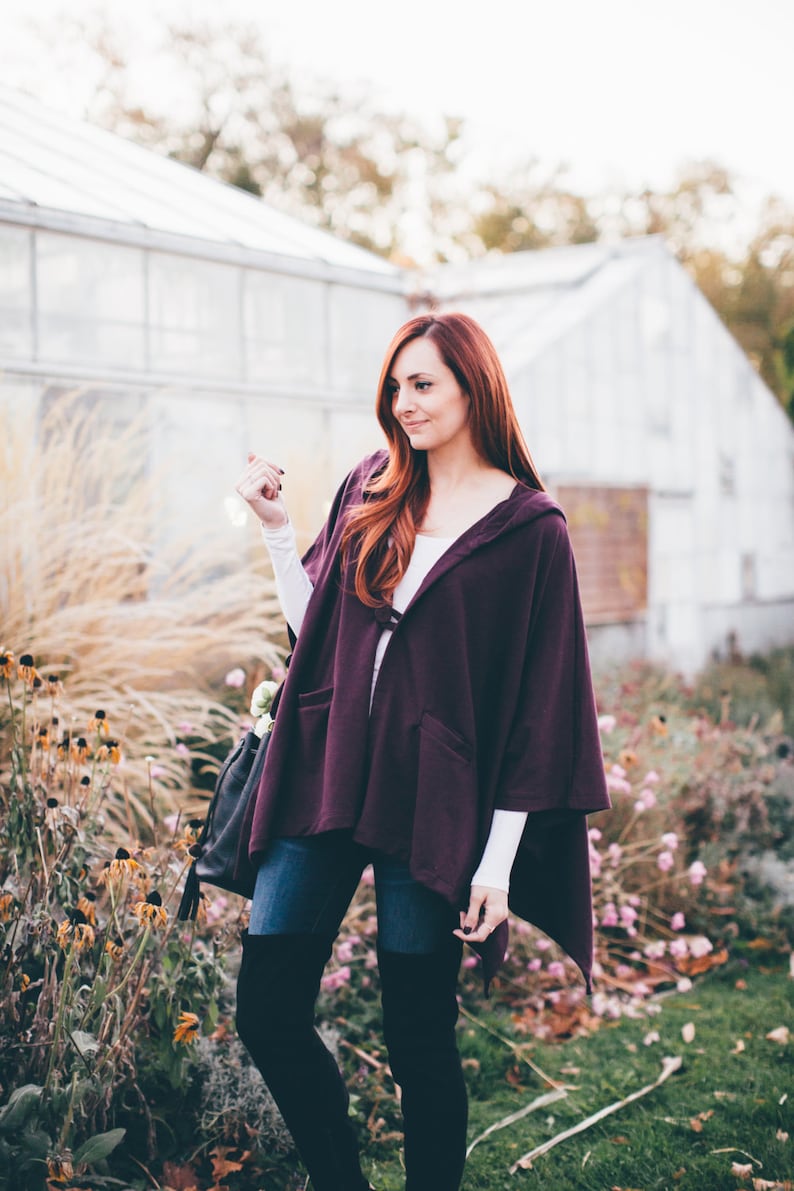 May include: A woman wearing a purple hooded cape, blue jeans, and black over-the-knee boots. She is standing in a garden with flowers and greenery.