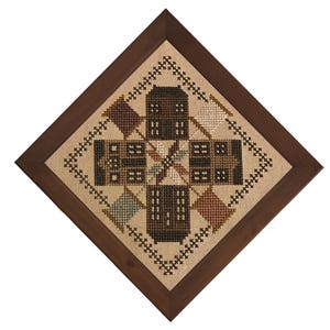 Op de afbeelding: Een ingelijste kruissteek sampler met een geometrisch ontwerp van gestileerde huizen en ruitvormen in bruin, beige en groen. Het kunstwerk is gevat in een donkerbruine houten lijst, wat een rustieke esthetiek creëert.