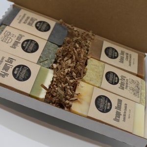 May include: A box containing six bars of handmade soap. The soaps are wrapped in brown paper with labels that list the scents: Key Lime Pie, Island Bay, Grapefruit Party, Orange Blossom, Lavender & Rosemary, and Herbal Mint. The soaps are nestled in brown paper shred.