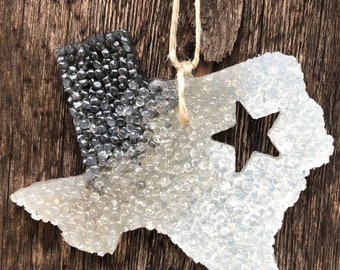 Puede incluir: Un ambientador de coche en forma de Texas negro y blanco con un recorte de estrella. El ambientador está hecho de un material transparente con pequeñas cuentas en todo. Está colgado de una cuerda.