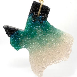 May include: A teal, green, and white Texas-shaped car air freshener with a black top. The air freshener is hanging from a string.