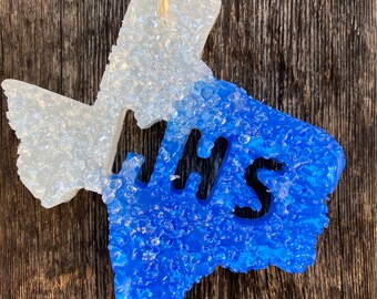 Puede incluir: Un ambientador con forma de Texas, dividido en secciones blancas y azules, con las letras "JHS" en negro. El ambientador está suspendido por una cuerda de color natural sobre un fondo de madera rústica.