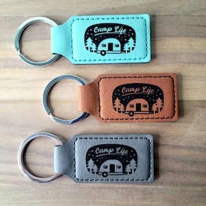 May include: Three leather keychains with silver key rings. Each keychain has a rectangular tag with a black and white design of a camper and trees. The text "Camp Life" is on each tag. The keychains are in turquoise, brown, and gray.