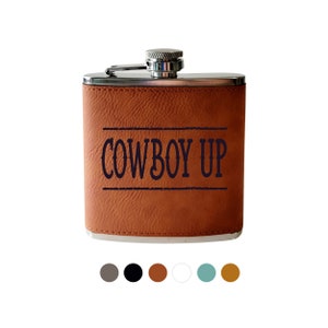 May include: A brown leather-wrapped flask with a silver cap and hinge. The text "COWBOY UP" is printed in dark blue. The flask is rectangular and has a stainless steel top.