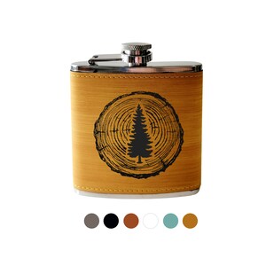 May include: A brown leather flask with a silver top and a black silhouette of a tree inside a circle on the front. The flask has a wood grain pattern.