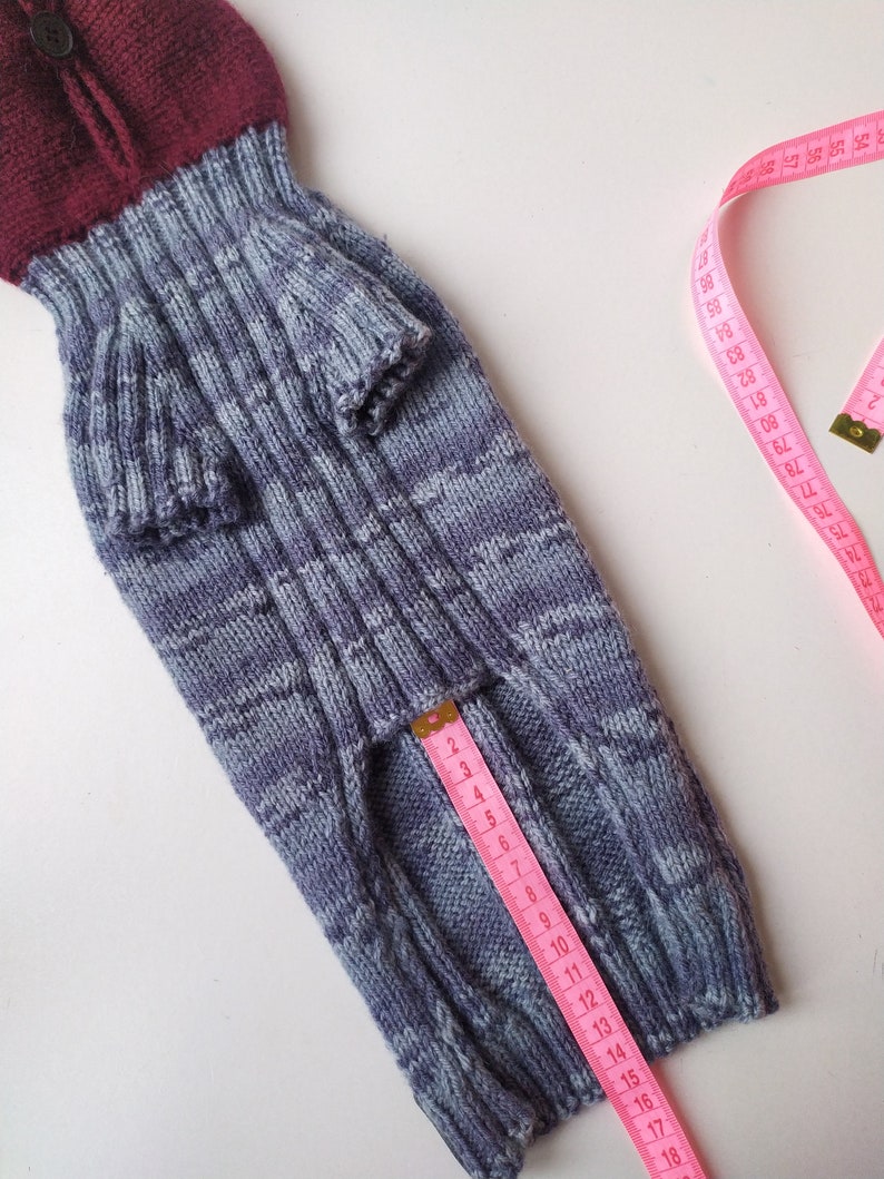 Może przedstawiać: Ręcznie robiony sweter dla psa w odcieniach niebieskiego i bordowego, zapinany na guziki. R&oacute;żowa miarka pokazuje pomiar około 25 cm. Sweter ma prążkowane detale i rękawy.