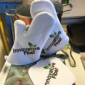 May include: Two white oven mitts with the text "Innovative Fiber" and a green plant graphic. A white pot holder with the same text and graphic is on a floral patterned surface.