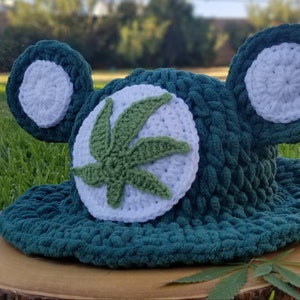 May include: A handmade, teal crochet hat with white accents and a green cannabis leaf design. The hat features two large, round ear-like shapes and is displayed on a wooden surface. A glass jar with dried buds is visible in the background.