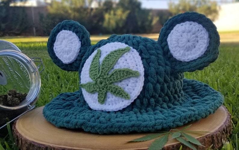 May include: A handmade, teal crochet hat with white accents and a green cannabis leaf design. The hat features two large, round ear-like shapes and is displayed on a wooden surface. A glass jar with dried buds is visible in the background.