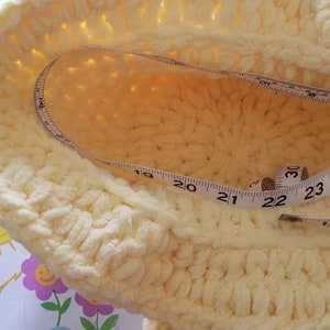May include: A light yellow, crocheted hat with a measuring tape inside. The tape shows measurements in inches, with the hat's diameter appearing to be approximately 22 inches. The hat is sitting on a colorful, patterned surface.