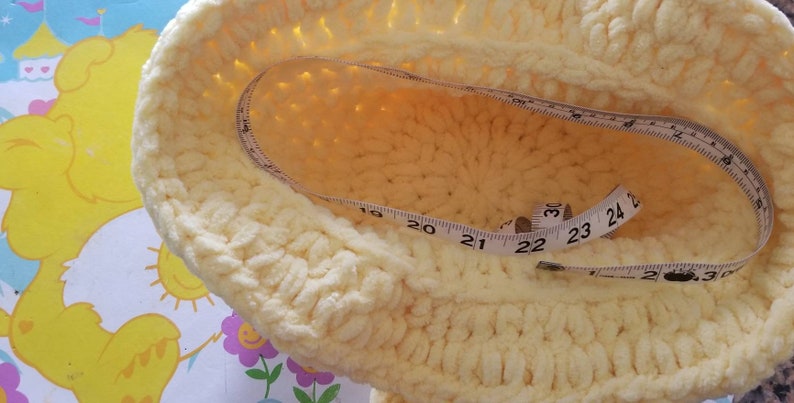 May include: A light yellow, crocheted hat with a measuring tape inside. The tape shows measurements in inches, with the hat's diameter appearing to be approximately 22 inches. The hat is sitting on a colorful, patterned surface.
