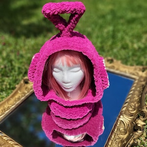 May include: A bright fuchsia crochet hood featuring a prominent triangular antenna on top. The textured, plush yarn construction covers the head and neck area, designed with a wide opening that frames the face, showcasing a vibrant pink color.