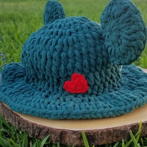 May include: A teal crochet hat with large, rounded ears and a red heart applique. The hat is made of a chunky yarn and sits on a wooden slice, with green grass in the background. The hat's design is reminiscent of a frog.