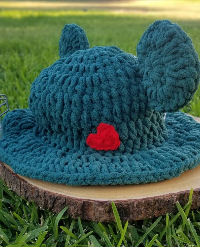 May include: A teal crochet hat with large, rounded ears and a red heart applique. The hat is made of a chunky yarn and sits on a wooden slice, with green grass in the background. The hat's design is reminiscent of a frog.