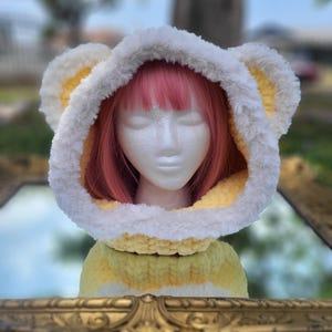 May include: A soft, chunky knit hood in pale yellow with a thick, white faux fur trim along the edge. The hood features two rounded, bear-like ears on top, crafted from the same yellow knit and white faux fur material, displayed on a mannequin.
