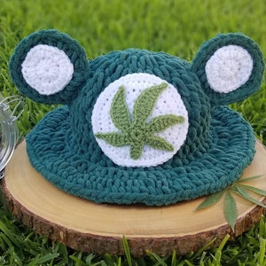 May include: A handmade, dark green crochet hat with white ear accents and a white circle featuring a green cannabis leaf. The hat is displayed on a wooden slice against a backdrop of green grass.