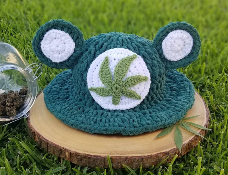 May include: A handmade, dark green crochet hat with white ear accents and a white circle featuring a green cannabis leaf. The hat is displayed on a wooden slice against a backdrop of green grass.