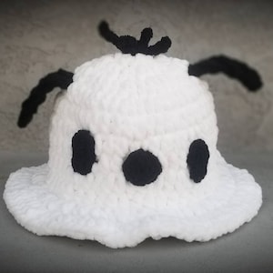Puede incluir: Sombrero blanco de ganchillo con detalles de hilo negro en forma de fantasma. El sombrero tiene un ala ancha y ojos negros.
