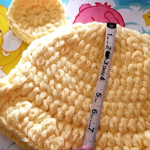 May include: A handmade, light yellow crochet hat with bear ears. The hat is measured with a tape measure, showing a height of approximately 6 inches. The background features a Care Bear illustration.