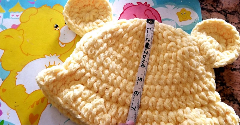 May include: A handmade, light yellow crochet hat with bear ears. The hat is measured with a tape measure, showing a height of approximately 6 inches. The background features a Care Bear illustration.