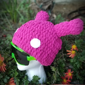 Puede incluir: Un gorro de punto rosa con un pompón blanco y unas gafas de esquí verde neón. El gorro tiene forma de conejo con orejas largas.