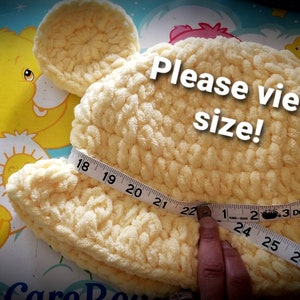 May include: A light yellow, handmade crochet hat shaped like a bear's head with ears. A measuring tape shows the hat's size. The text "Please view size!" is overlaid on the image. The hat measures approximately 22 inches in diameter.