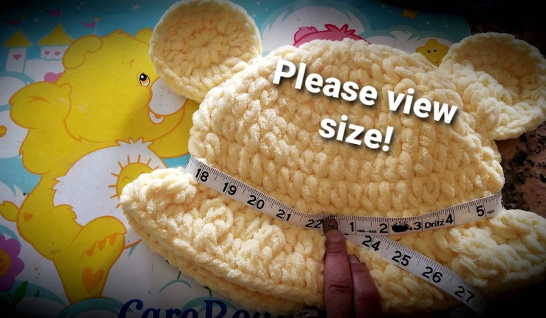 May include: A light yellow, handmade crochet hat shaped like a bear's head with ears. A measuring tape shows the hat's size. The text "Please view size!" is overlaid on the image. The hat measures approximately 22 inches in diameter.