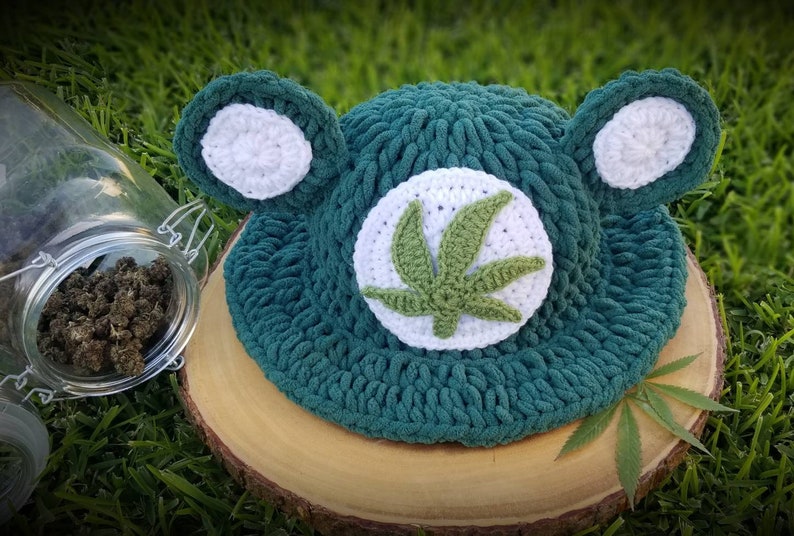 May include: A handmade, teal crochet hat with white ear accents and a white circle featuring a green cannabis leaf. The hat is displayed on a wooden slice, with a jar of cannabis buds in the background.