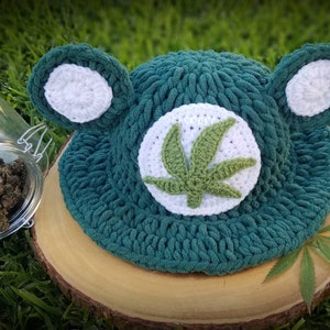 May include: A handmade, teal crochet hat with white ear accents and a white circle featuring a green cannabis leaf. The hat is displayed on a wooden slice, with a jar of cannabis buds in the background.