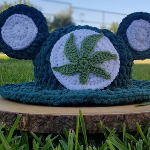 May include: A handmade, dark teal crochet hat with white accents and a green cannabis leaf design. The hat features large, round ear-like embellishments. The hat is displayed on a wooden slice, with a glass jar and greenery in the background.