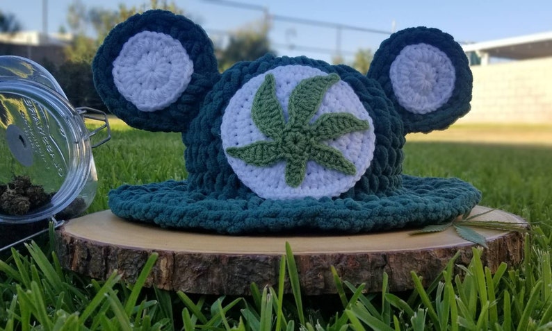 May include: A handmade, dark teal crochet hat with white accents and a green cannabis leaf design. The hat features large, round ear-like embellishments. The hat is displayed on a wooden slice, with a glass jar and greenery in the background.