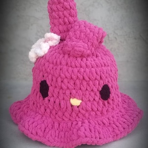 Puede incluir: Un sombrero de cubo de crochet rosa con un lazo de crochet blanco y ojos bordados negros y amarillos.