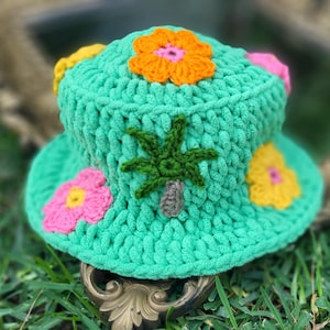 Könnte beinhalten: Ein grüner Häkel-Bucket-Hut mit bunten Häkelblumen und einem Palmenmotiv.