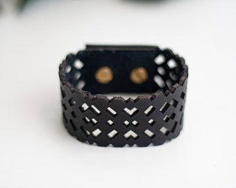 Puede incluir: Un brazalete de cuero negro con un diseño de corte geométrico. El brazalete tiene dos broches de metal dorado.