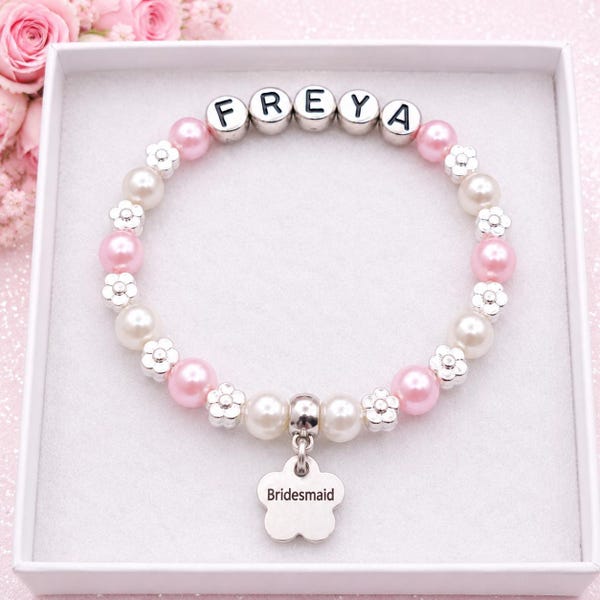 May include: A beaded bracelet featuring alternating pink and white pearls, silver-toned flower spacers, and letter beads spelling FREYA. A silver-toned flower-shaped charm hangs from the center, engraved with the word Bridesmaid.