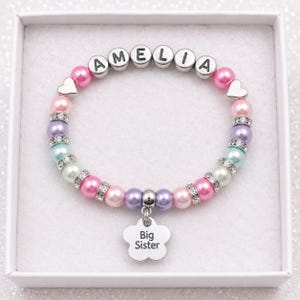 May include: A beaded bracelet featuring pastel-coloured pearls, silver-toned spacers with embedded crystals, and letter beads spelling AMELIA. A silver-toned flower-shaped charm hangs from the centre, engraved with the text Big Sister.