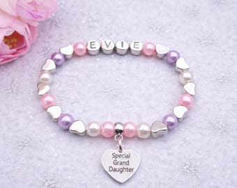 May include: A beaded bracelet featuring alternating light pink, white, and lavender pearls interspersed with silver-toned heart-shaped beads. The centre displays silver letter beads spelling EVIE, and a heart-shaped charm engraved with Special Grand Daughter.