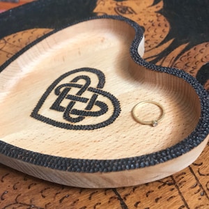 May include: A heart-shaped wooden trinket tray with a Celtic knot design burned into the wood. The tray has a black beaded border and holds a small gold ring with a single clear stone.