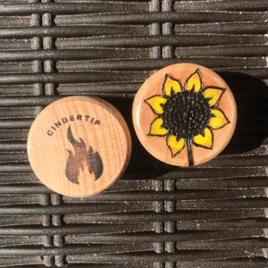 May include: Two circular wooden plugs with flat faces. One plug features a black wood-burned flame graphic with the text CINDERTIP above it. The other plug displays a wood-burned sunflower design with yellow-painted petals and a dark centre.