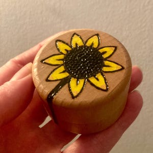 May include: A small, round, light-coloured wooden container with a lid. The top of the lid features a hand-drawn sunflower design with a dark brown centre and bright yellow petals, outlined in black ink that extends down the side of the container.