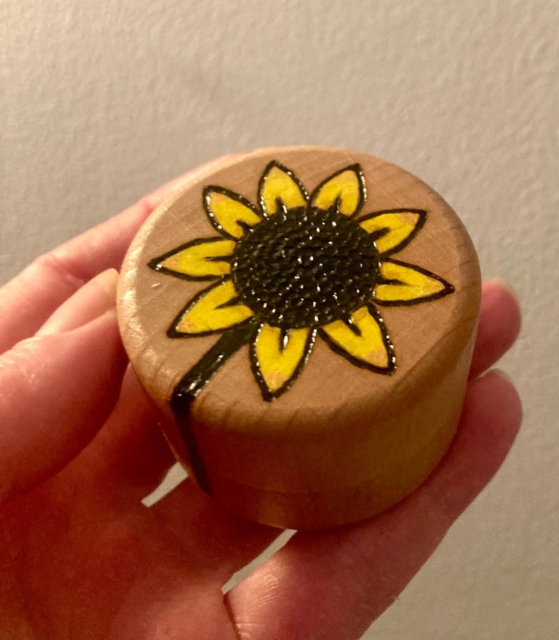 May include: A small, round, light-coloured wooden container with a lid. The top of the lid features a hand-drawn sunflower design with a dark brown centre and bright yellow petals, outlined in black ink that extends down the side of the container.