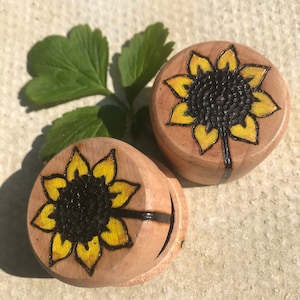 May include: Two round wooden plugs featuring a wood-burned sunflower design with yellow-painted petals and a dark brown textured centre. The sunflower artwork is detailed with black outlines, and the natural light wood grain is visible on the surface.