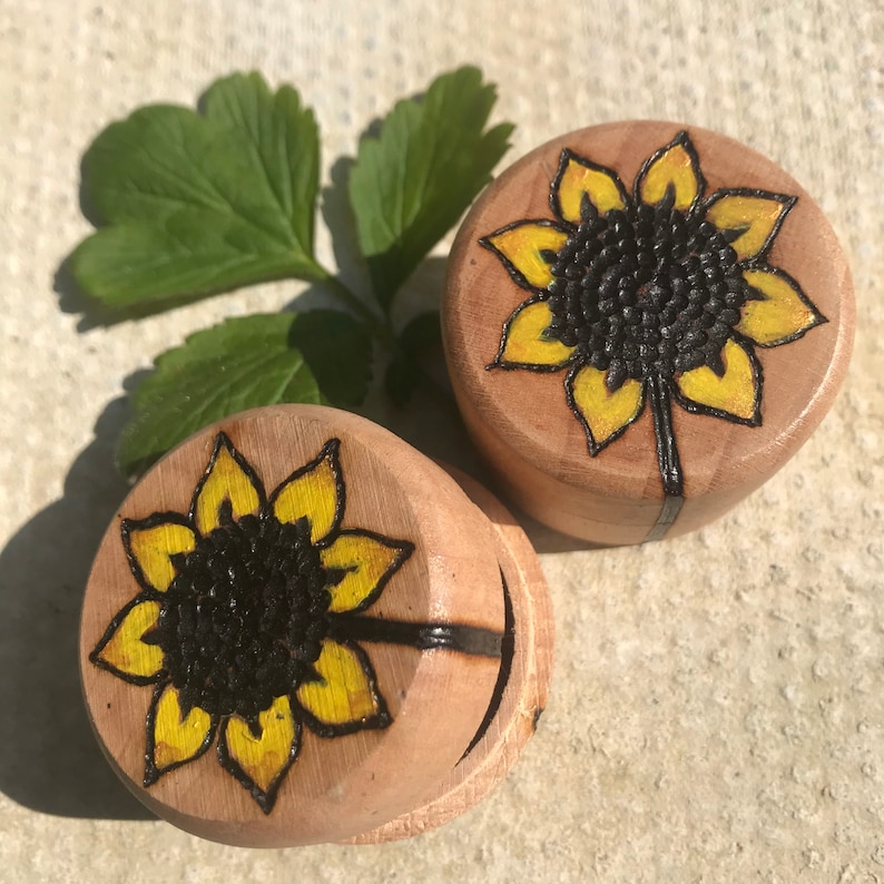 May include: Two round wooden plugs featuring a wood-burned sunflower design with yellow-painted petals and a dark brown textured centre. The sunflower artwork is detailed with black outlines, and the natural light wood grain is visible on the surface.