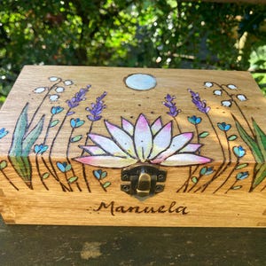 Peut inclure: Une boîte en bois avec un motif floral peint sur le couvercle. Le motif représente un lotus blanc avec des accents roses, entouré de feuilles vertes et de fleurs de lavande violettes. La boîte a un loquet en laiton et le nom "Manuela" est gravé sur le devant.